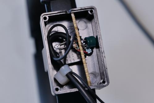 Een klein electronicaprintje met enkele onderdelen en 2 draden in een klein zwart plastic kastje, gemonteerd aan een koptelefoon