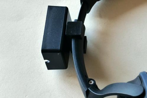 Een klein plastic zwart kastje dat met een 3d-geprint C-houdertje om een koptelefoon geschoven wordt