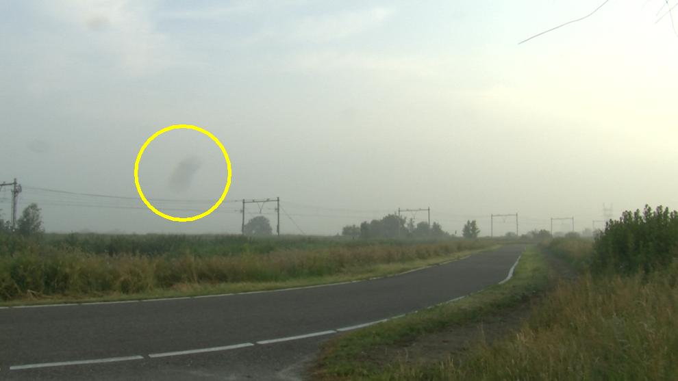 Een video-still van een zomerse ochtendlucht, fietspad op de voorgrond, spoorweg op de achtergrond en een donkere vlek in het beeld door een stofje op de lens aan de binnenkant van een videocamera
