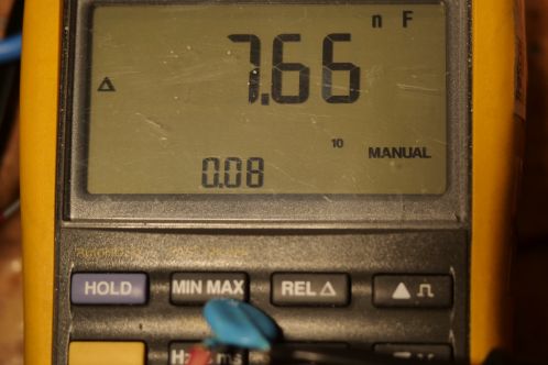 Een geel met grijze multimeter die op het zwart-witte LCD scherm 7,66 nanoFarad toont, de waarde van een van de uitgenomen hoogspanningscondensators na vervanging