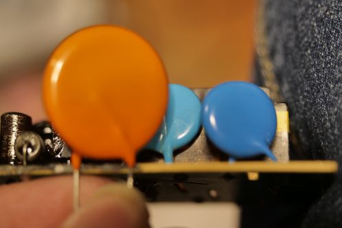 Drie verschillende schijfvormige hoogspanningscondensatoren, 1 oranje en twee blauwe, waarvan 1 blauwe gesoldeerd in een elektronicaprint