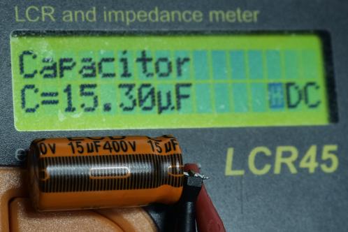 Een kleine, oranje-zwarte 15 uF 400 Volt elco aangesloten op een meetapparaat dat op een groen LCD scherm 15,30 microfarad toont