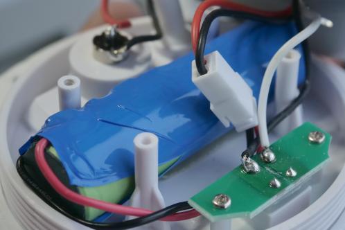 Een lithium-cel met gedeeltelijk gescheurde blauwe krimpkous, aangesoldeerde draden verbonden met een connector, gemonteerd op een witte kunststof plaat, de licht-eenheid van een Le Zen Lux lamp