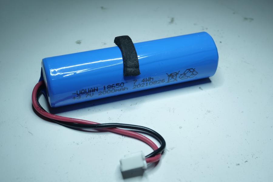 Een blauwe cilindrische lithium-cel in 18650 formaat met kabeltje met rode en zwarte draad met connector, de accu uit een Le Zen Lux lamp