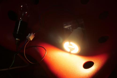 Een rode kruk waarop de gerepareerde Osram AC01754 LEDlamp zonder transparante bol ligt, in ingeschakelde toestand en daarbij een enorme bak licht verspreidt