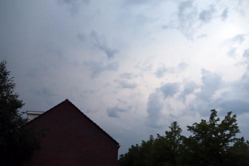 Boomtoppen en een puntdak van een huis met daarboven woeste bewolking waarbij het daglicht rap afneemt bij nadering van een stevige onweersbui