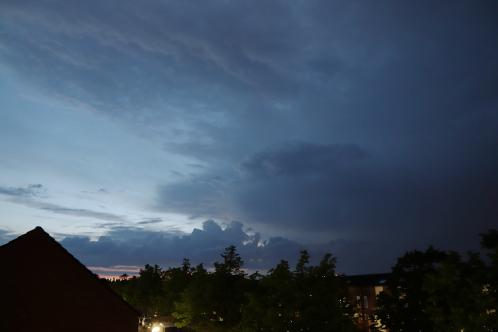 Boomtoppen en een puntdak met links een heldere avondlucht en rechts een wegtrekkende onweersbui