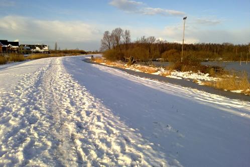 Bewolking in de verte maar de zon schijnt op witte, door sneeuw bedekte velden en fietspad bij Ringvaart in het Twiske, terwijl bomen en struiken vrijwel volledig sneeuwvrij zijn, na sneeuwjacht op 11 december 2017, foto genomen op 12 december 2017